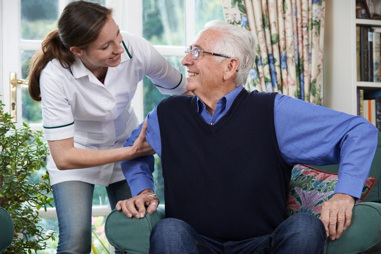 A caregiver helping a smiling senior gentleman up from his armchair